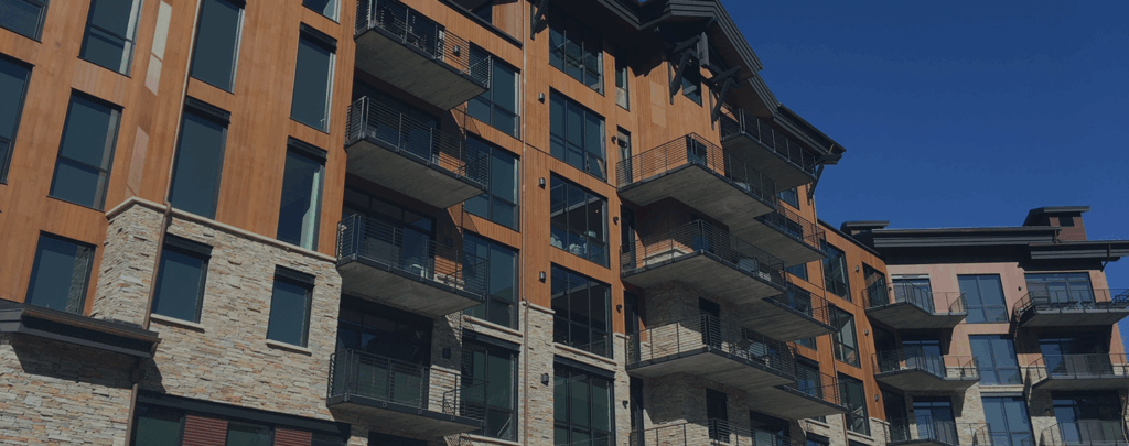 Large modern residential building with Shakertown® vertical cedar panel siding, stone accents, multiple balconies, and large windows under a clear blue sky banner header